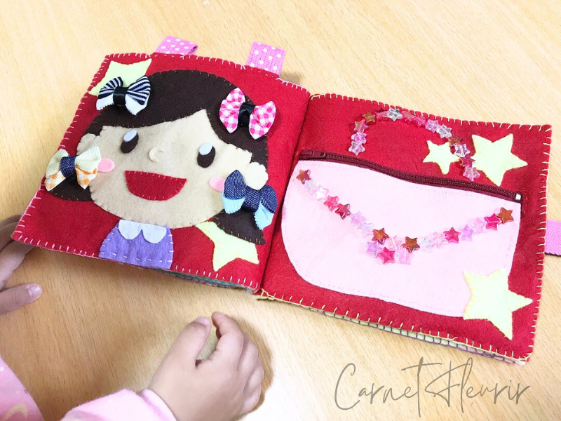 布絵本のページで、女の子の髪にリボンをつけ外しできる仕掛けの様子、左はフェルト製のバッグの仕掛け。開け閉めできるポケットで、小さなアイテムを入れられる工夫がされている。