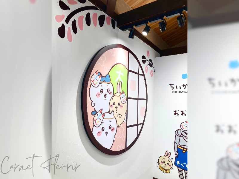 階段を上がって横をみると飾られている、ちいかわ・ハチワレ・うさぎの絵。頭には狐面をつけている。