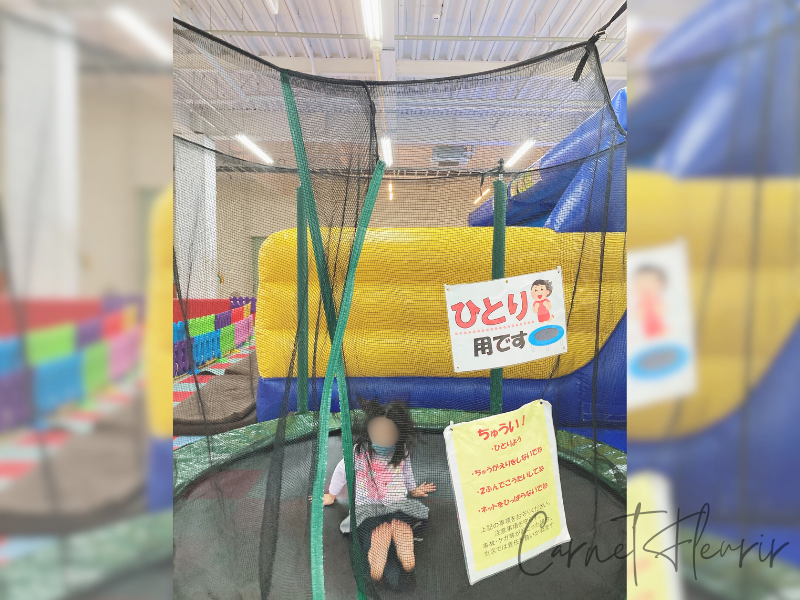 ネットで囲まれた一人用トランポリンで遊ぶ女の子。後ろにはカラフルな遊具や注意書きの看板が見える