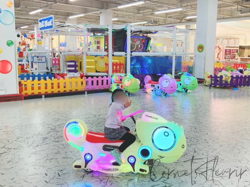 光るカメレオン型の乗り物に乗る子ども。カラフルなライトと室内遊び場のにぎやかな雰囲気が特徴的。