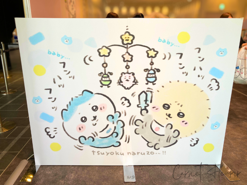 ハチワレとラッコが天井のモビールを見上げて遊んでいるイラストパネル。ベビーカラーの飾りや「tsuyoku naruzo…!!」のセリフも可愛い。
